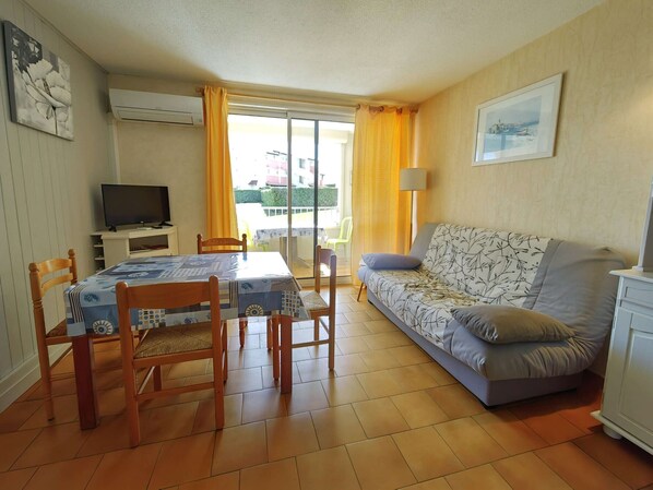 A living room with a sofa, a dining table with chairs, a television, and a balcony with yellow curtains.