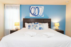 Ein ordentlich bezogenes Bett mit weißen Leinen und zwei gefalteten Handtüchern, ein blaues abstraktes Gemälde an der Wand und zwei Nachttischlampen auf Holz-Nachttischen.