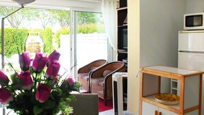A room with a sliding glass door leading to a garden, a kitchenette with a microwave, a small table with a bowl, and a chair.