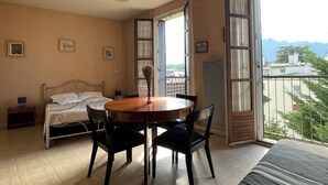 A bedroom with a bed, a dining table with chairs, a small bedside table, a vase with dried flowers, and a large window with a view of buildings and mountains. - Spacious city-center studio, pets allowed (Aix-les-Bains)