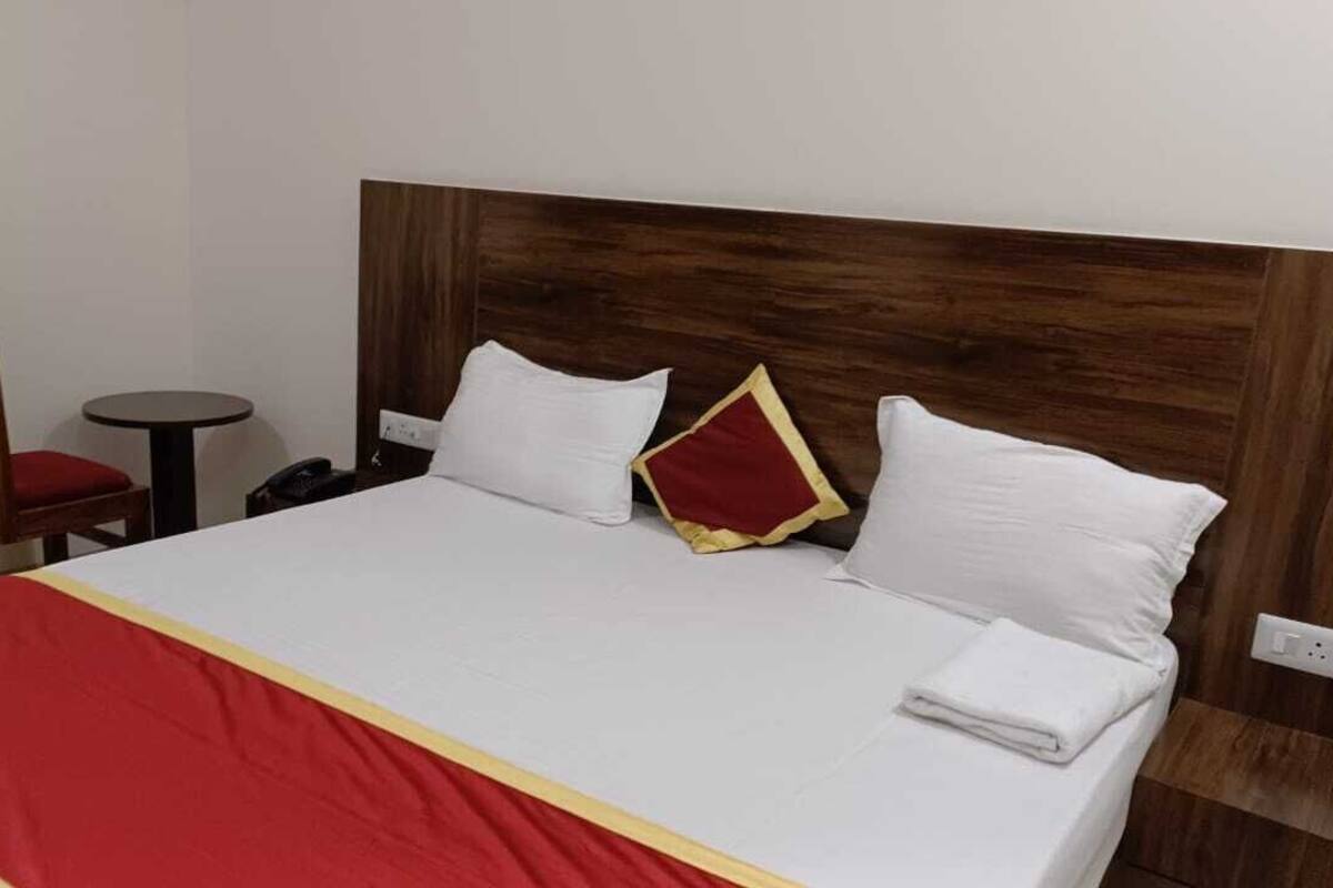 Une chambre d’hôtel avec un lit, une tête de lit en bois, une couvre-lit rouge et blanche, et une petite table ronde avec une chaise.