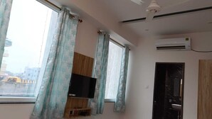 Ein Zimmer mit an einer Holzvertäfelung befestigtem Fernseher, zwei Vorhängen und einem Klimagerät, sowie einer Tür mit schwarzem Rahmen.