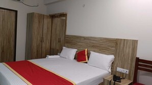Ein ordentlich bezogenes Bett mit einer roten und weißen Tagesdecke, zwei weißen Kissen und einem roten Zierkissen. Ein Holz-Kopfteil und ein Nachttisch mit Telefon und Buch neben dem Bett.