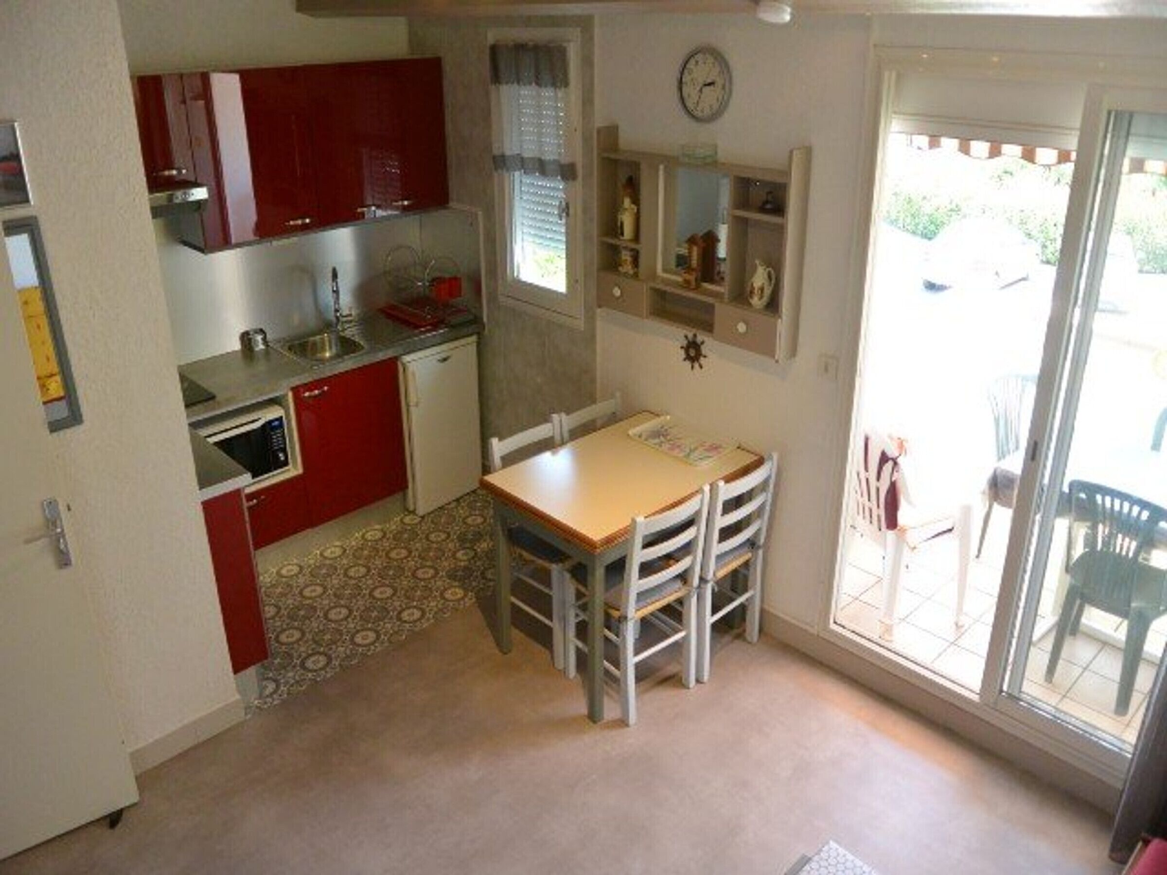 A compact kitchen with red cabinets, a microwave, and a sink. There's a dining table with chairs and a window with blinds.