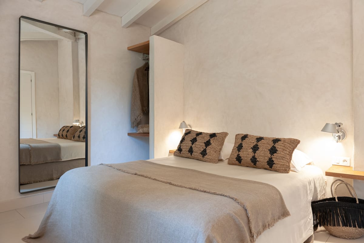 A bedroom with a bed, two pillows, a bedside lamp, a mirror, a hanging basket, and a wooden shelf.