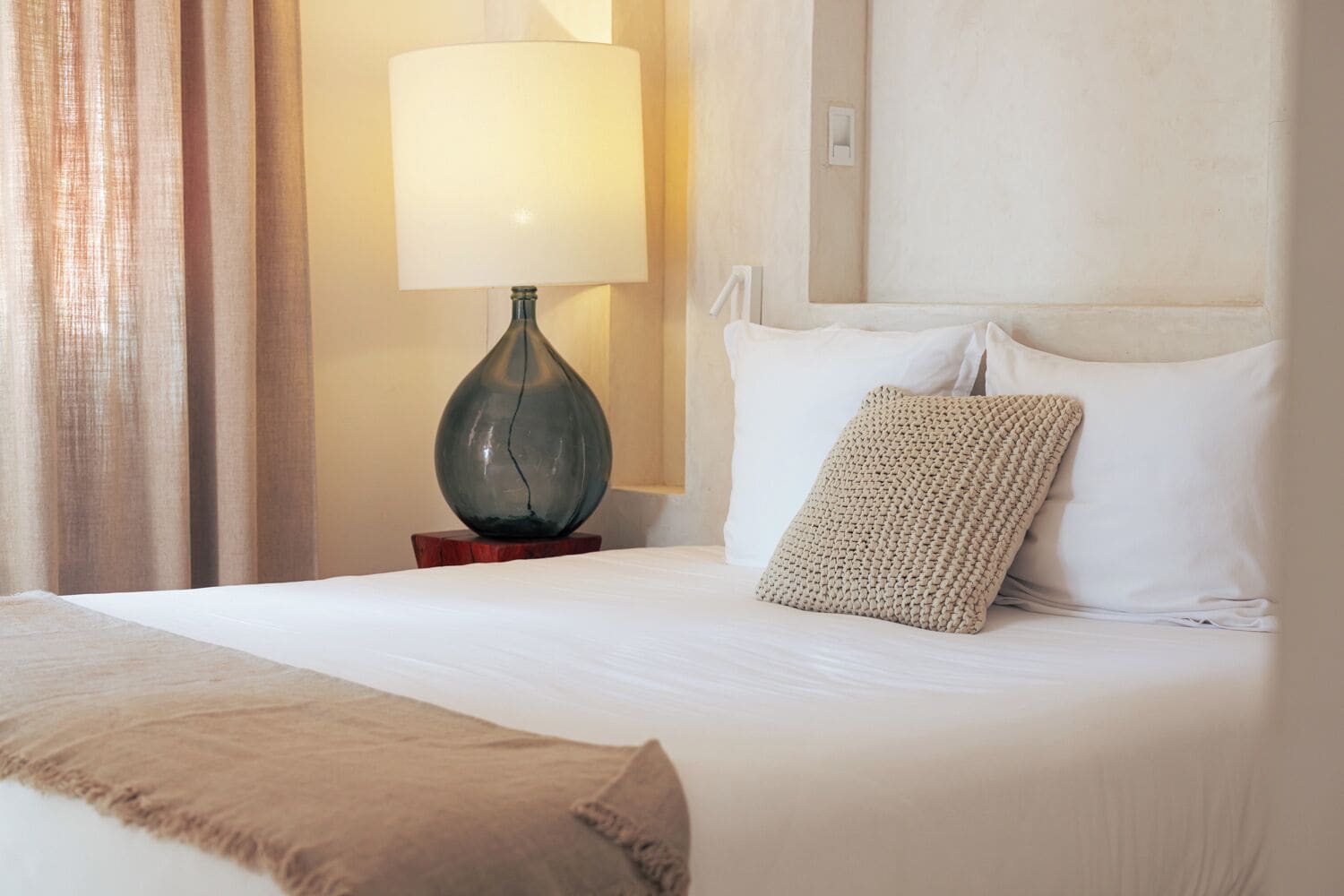 A neatly made bed with white linens, a beige throw blanket, and a woven throw pillow. A lamp with a green shade is on a wooden nightstand beside the bed.