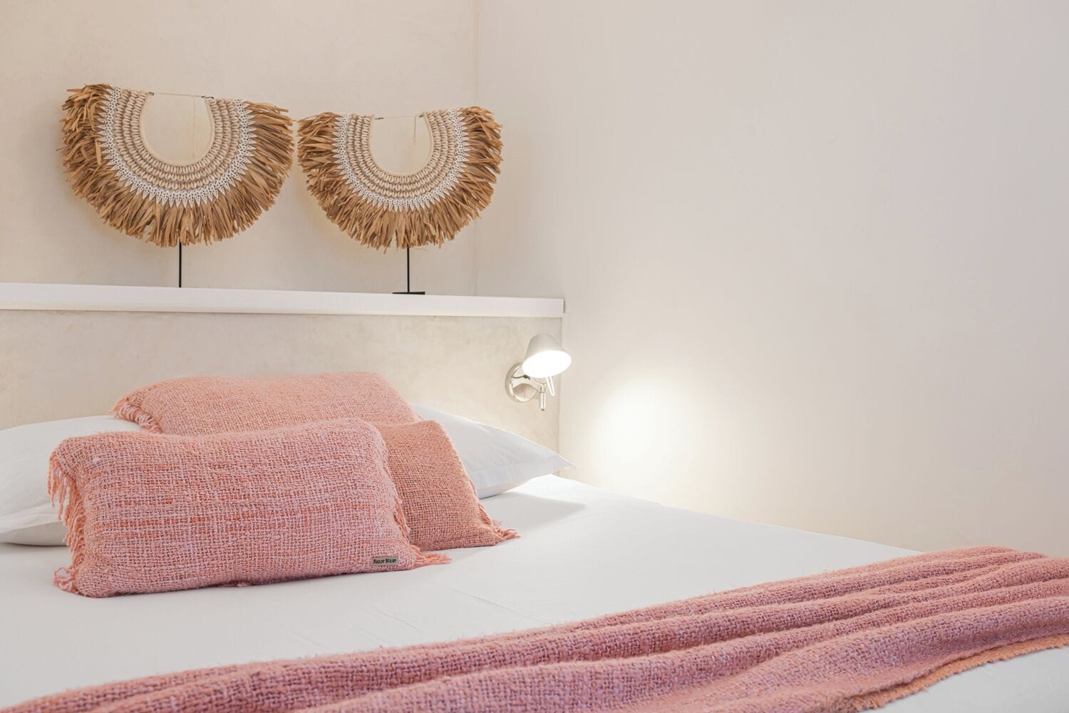 Una cama bien tendida con almohadillas rosadas con textura y una colcha, una lámpara de pared y dos obras de arte con flecos.