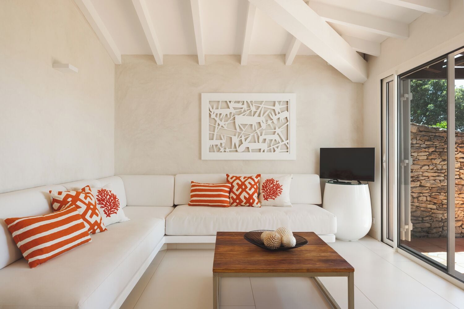 Una sala de estar moderna con un sofá blanco, almohadones decorativos, una mesa de centro de madera, un televisor de pantalla plana y un amplio ventanal con vistas a una pared de piedra.