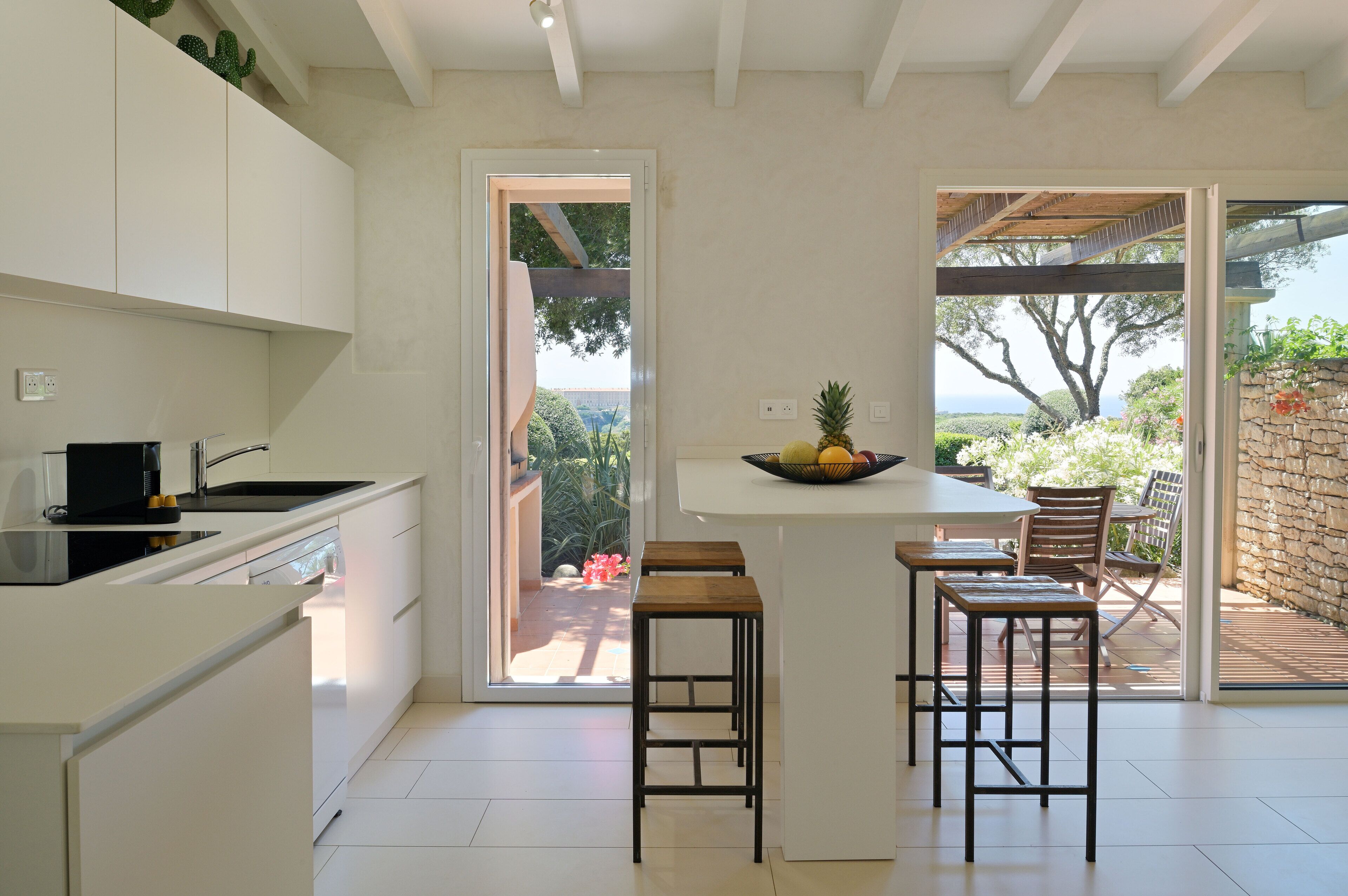 A modern kitchen with white cabinets, a central island with stools, and a view of the outdoors through a glass door.