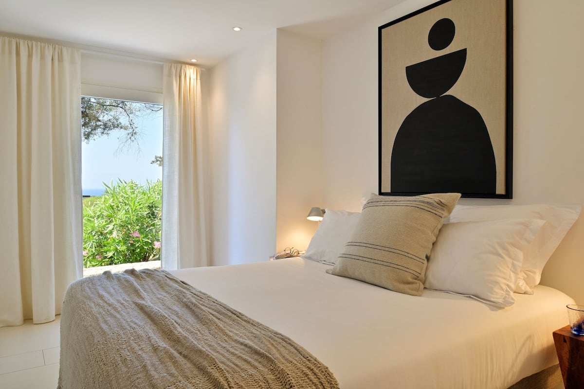 A bedroom with a bed, a nightstand, a lamp, and a large window with curtains.