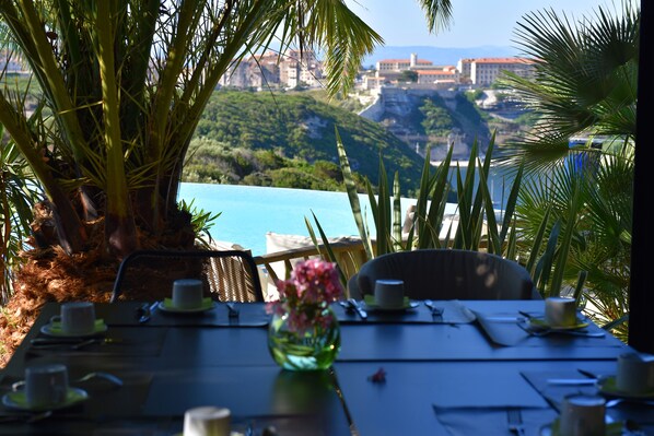 A table set for dining with a view of a coastal landscape, including buildings and greenery. - Résidence Terra Marina (Bonifacio)
