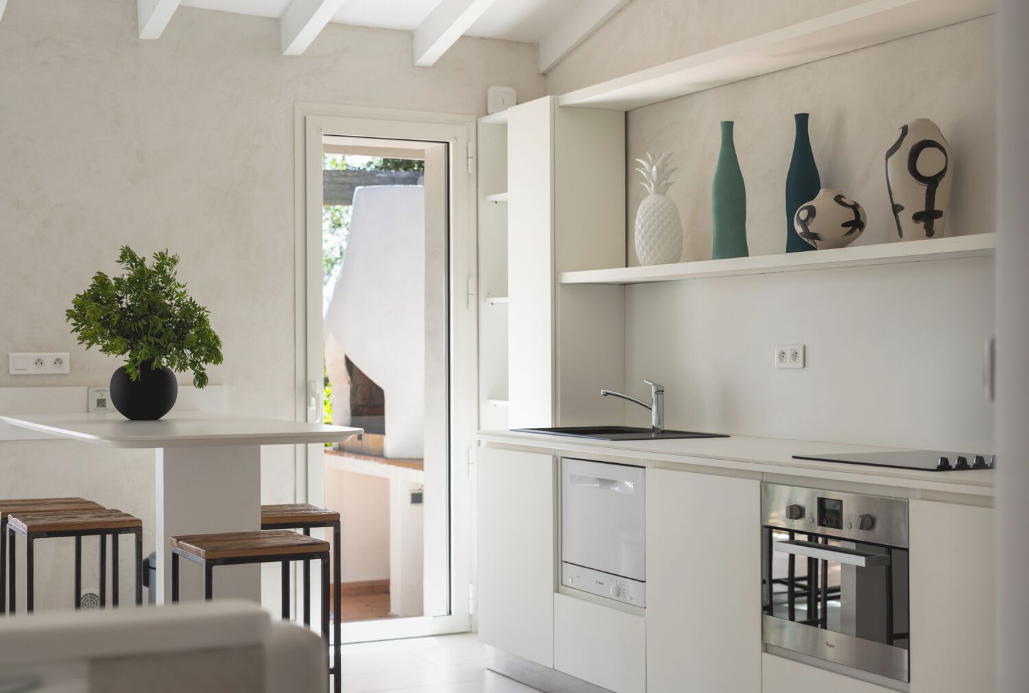 Una cocina moderna con armarios blancos, horno empotrado y un lavamanos. Hay una planta en maceta sobre una pequeña mesa y un estante con objetos decorativos.