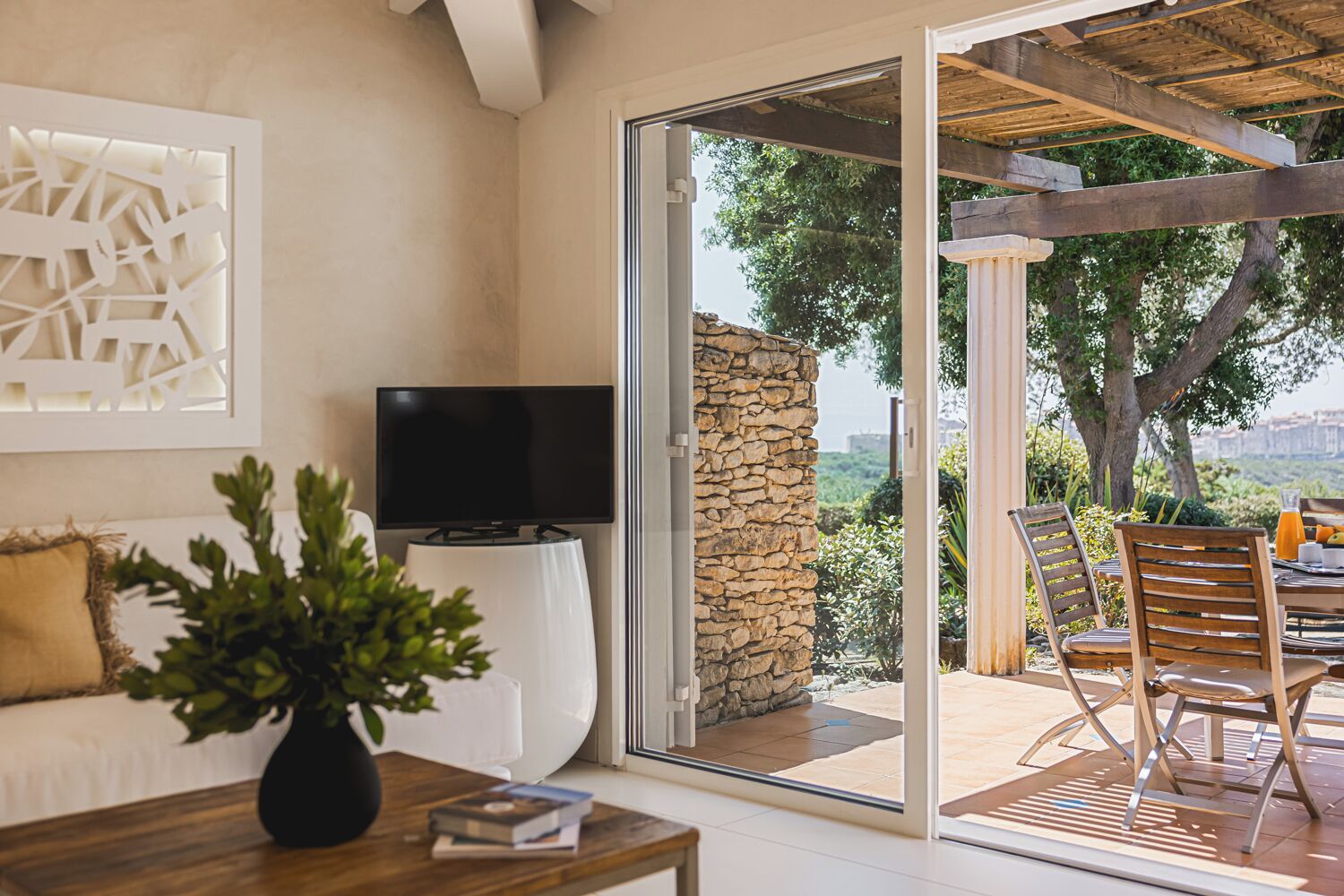Un salon moderne avec un téléviseur, un canapé blanc, une table basse avec des livres et une vue sur une terrasse extérieure dotée d’un mur en pierre et d’une pergola en bois.