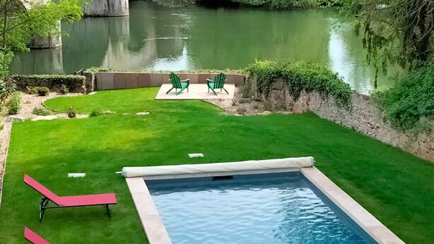 A rectangular swimming pool with a small deck area, surrounded by a well-maintained lawn and a stone wall.