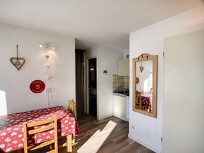 A small, cozy room with a dining table and chairs, a mirror, and a kitchen visible through an open door.