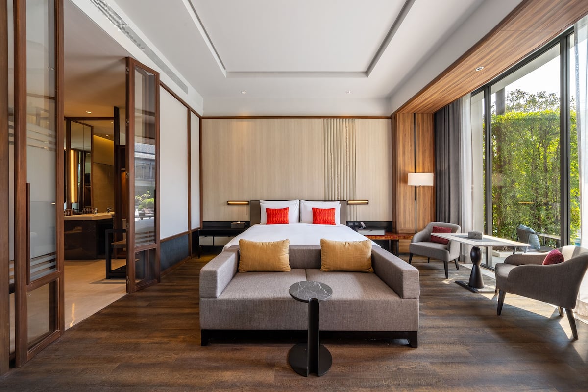 A modern hotel room with a large bed, a sofa, a small table, and a view of greenery through large windows.