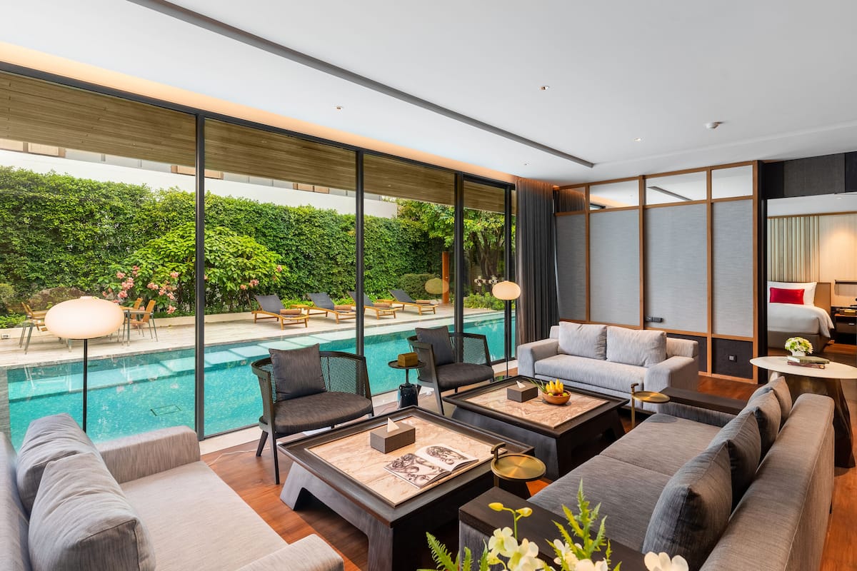 A modern hotel room with a large window overlooking a swimming pool and lush greenery. There are multiple seating areas with sofas and armchairs, and a small table with a lamp.