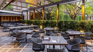 Een terras met meerdere tafels en stoelen, een pergola en een uitzicht op een gebouw op de achtergrond.