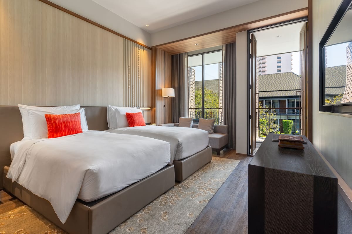 A modern hotel room with two beds, a small seating area, a balcony, and a view of the city.