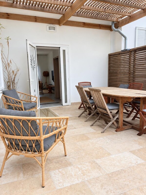 An outdoor patio with a wooden table and chairs, a wicker chair, and a door leading to an interior space.