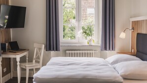 Un dormitorio con cama, escritorio con laptop, silla, televisor y ventana con cortinas.