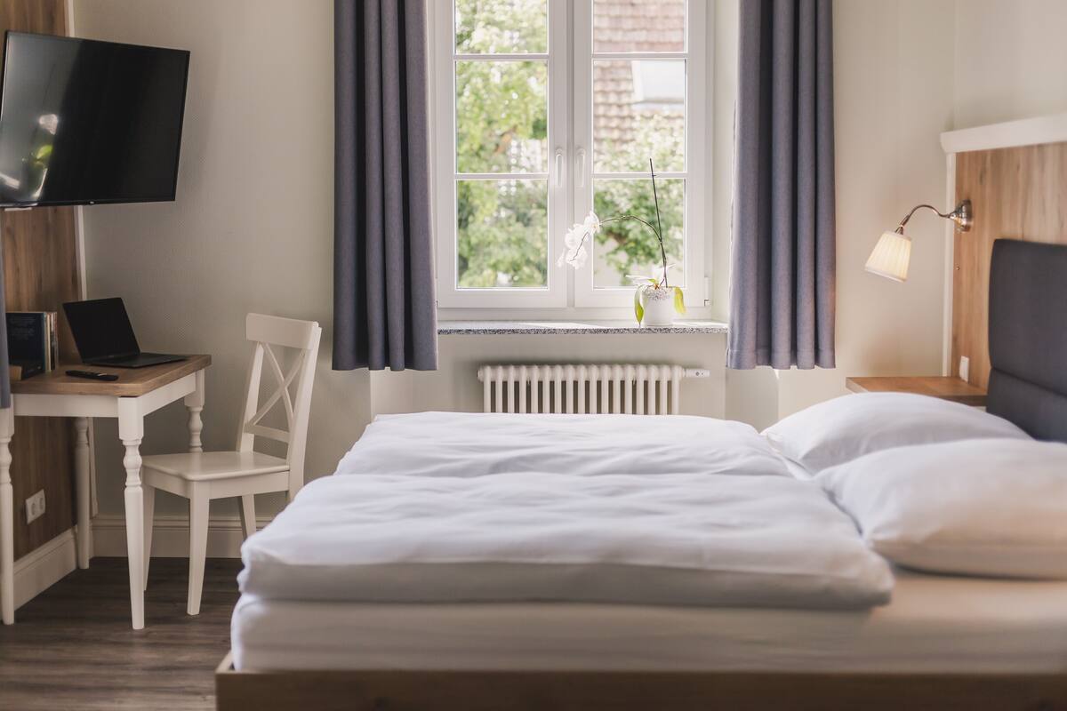 Een slaapkamer met een bed, een bureau met een laptop, een stoel, een televisie en een raam met gordijnen.