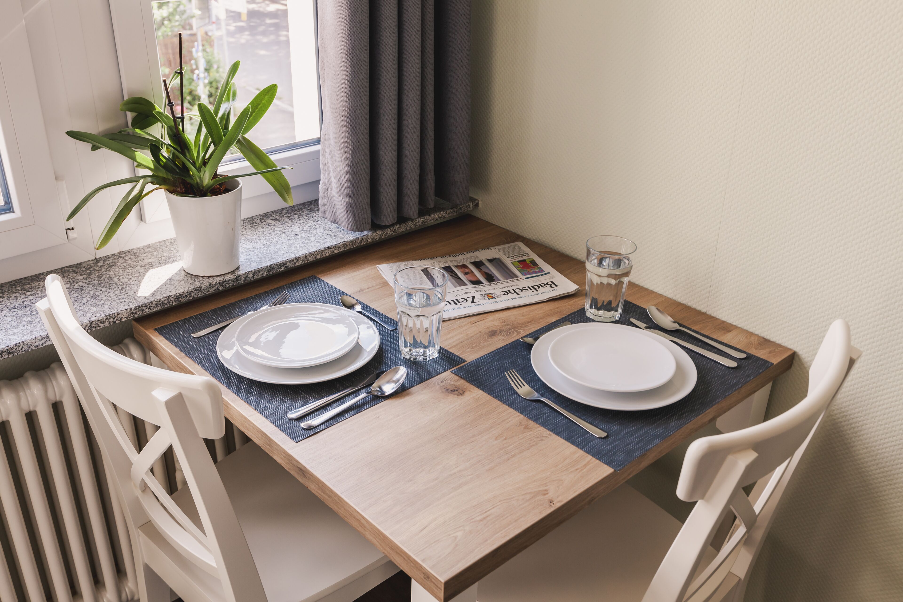 Een eettafel gedekt met borden, bestek en een glas water, met een plant en gordijnen op de achtergrond.