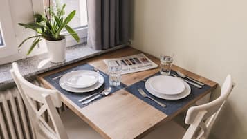 Una mesa de comedor dispuesta con platos, cubiertos y un vaso de agua, con una planta y cortinas al fondo.