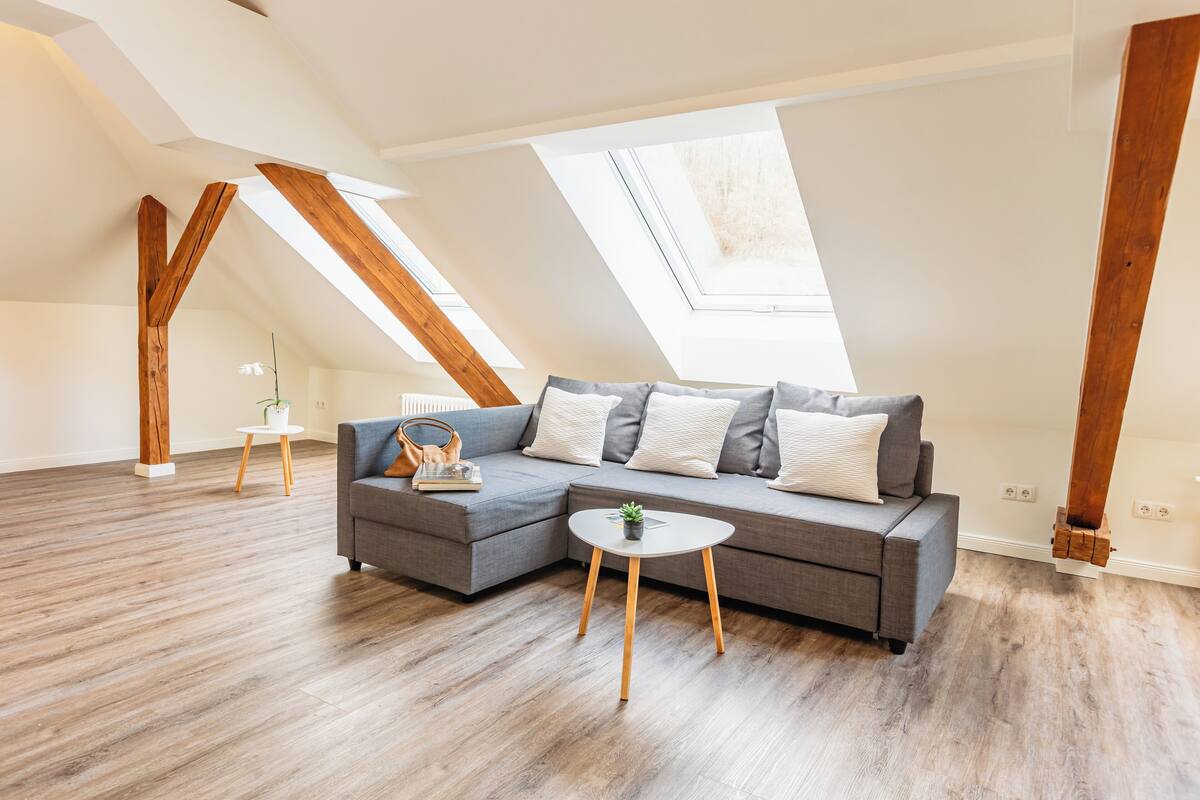 Een moderne loftkamer met een grijze modulaire bank, een klein rond tafeltje en houten balken.