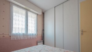 A bedroom with a bed, a window with lace curtains, a radiator, and a wardrobe. - Cozy Apartment Near Beach and Shops, with Balcony and Modern Amenities (Les Sables-d'Olonne)