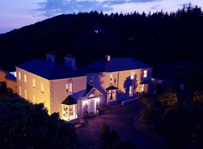A large, multi-story house with a well-lit entrance and a driveway leading to the front door.