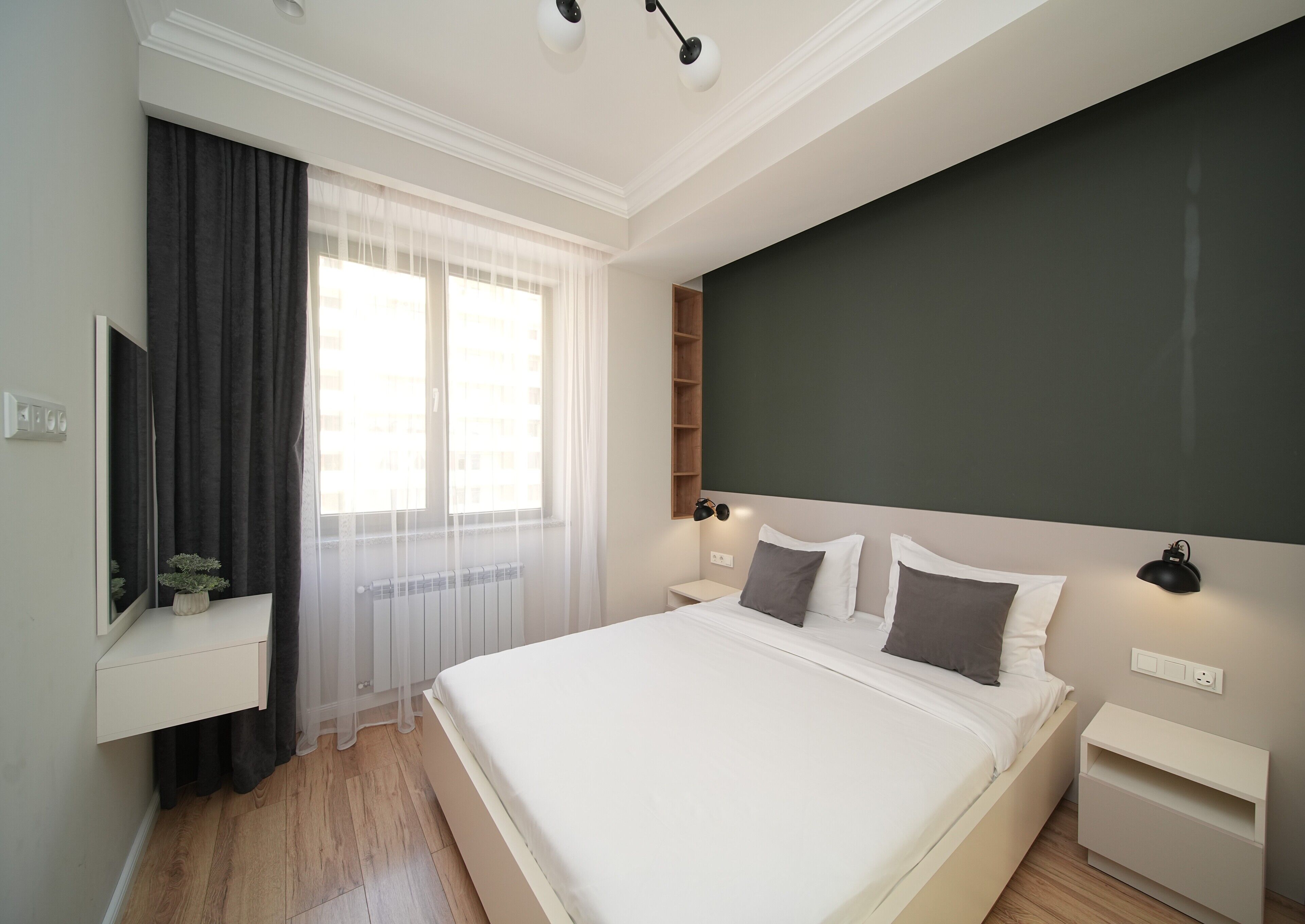 A modern bedroom with a large bed, a bedside table, a nightstand, a wall-mounted shelf, and a window with sheer curtains.
