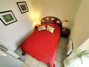 A neatly made bed with a red bedspread and two pillows, a wooden headboard, a bedside table with a lamp, and a framed picture on the wall. - Charming T2 with Terrace and Sea View 50m from Beach, Residence with Pool (Cap d'Agde)