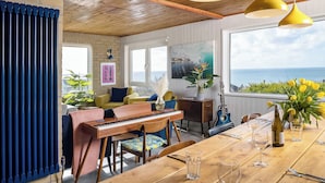 A cozy living room with a piano, a dining table set for two, and a view of the sea. - Sunset Beach House, Porthleven, with panoramic sea views. Comfortably sleeps 10 (Porthleven)