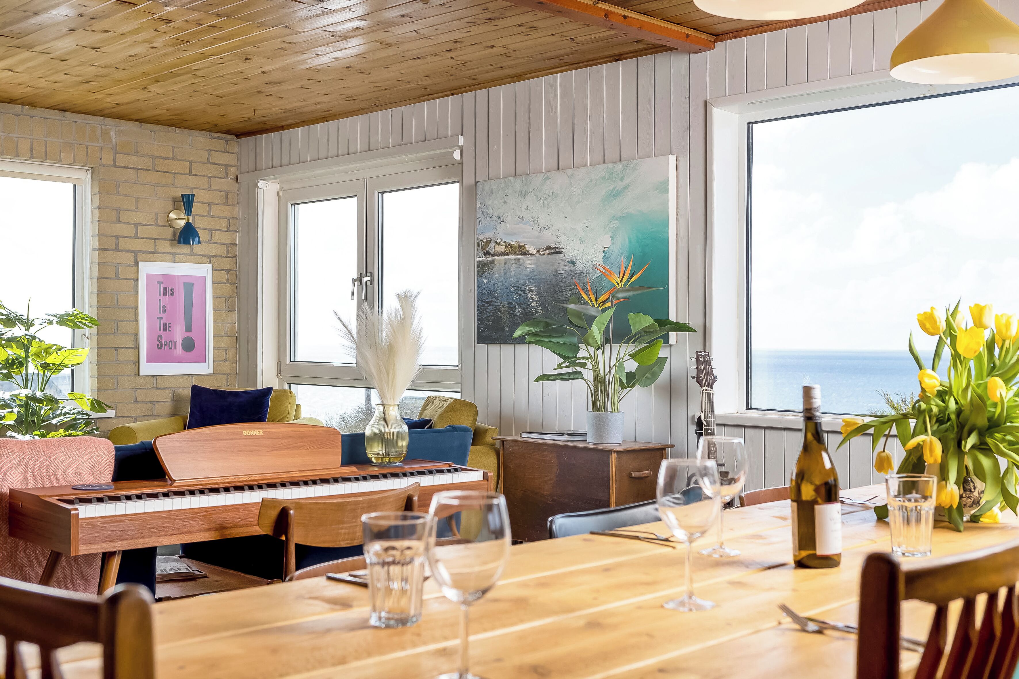 A dining area with a wooden table, chairs, a piano, and a view of the ocean through large windows.