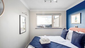 A neatly made bed with a blue quilt, white pillows, and a folded towel. There is a window with a view of the outside, a framed picture on the wall, and a ceiling light fixture. - Sunset Beach House, Porthleven, with panoramic sea views. Comfortably sleeps 10 (Porthleven)