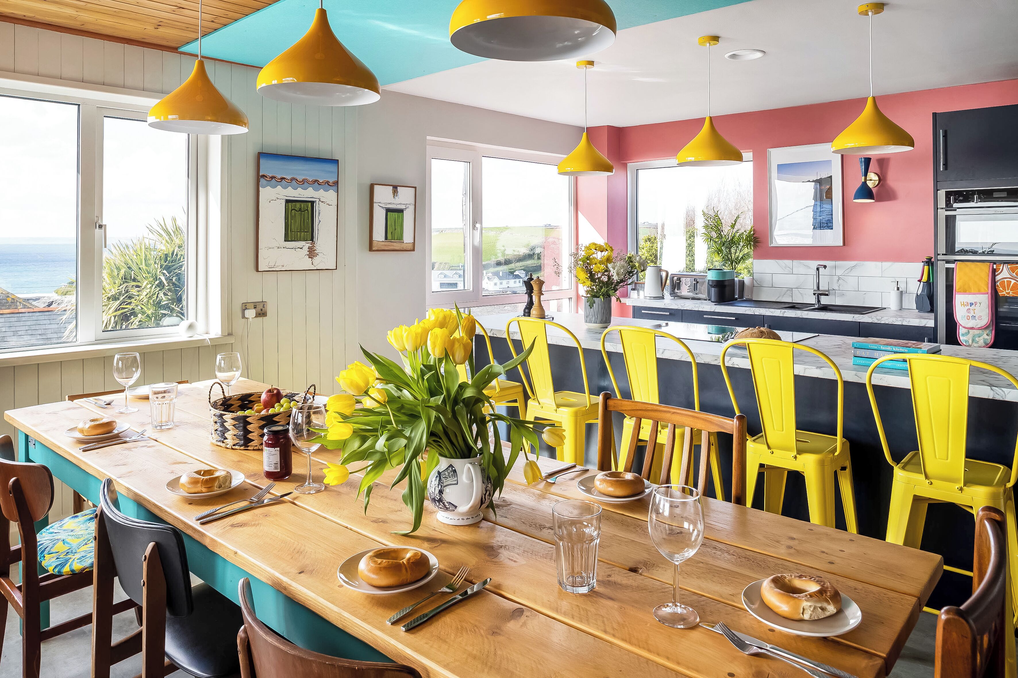 Een eetgedeelte met een houten tafel voor vier personen, gele stoelen en uitzicht op de oceaan door grote ramen.