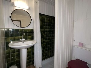 Un baño con un lavamanos blanco, un espejo redondo, paredes con azulejos verdes, una cortina blanca y un inodoro rojo.