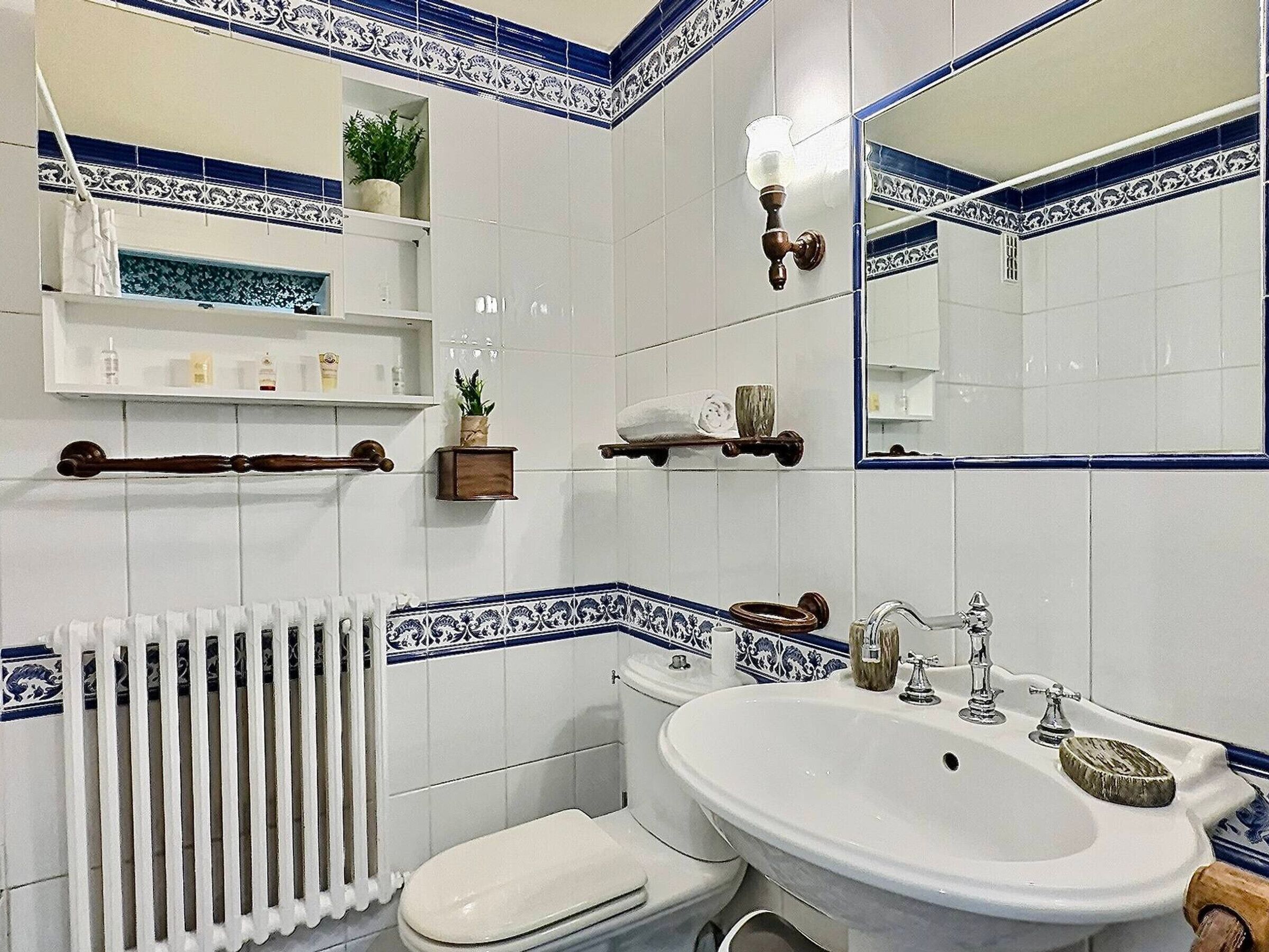 Un bagno con il water, un lavandino e uno specchio. Si vedono scaffali con articoli da toilette e una pianta.