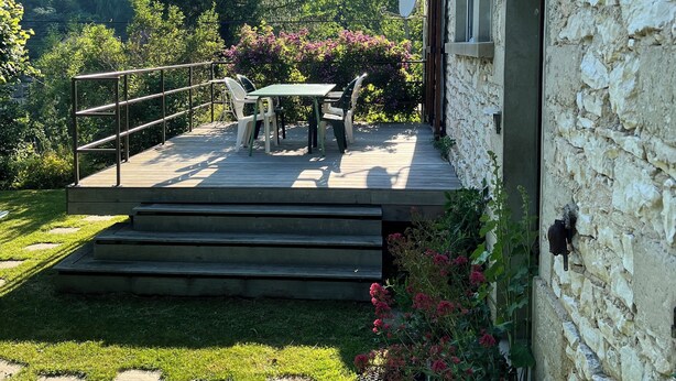 Een stenen gebouw met terras, buitenzitplaatsen en uitzicht op een beboste heuvel.