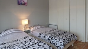 A bedroom with two beds, a nightstand, a lamp, a radiator, and a wardrobe. - Bright T2 with Balcony near the Thermal Baths (Balaruc-les-Bains)