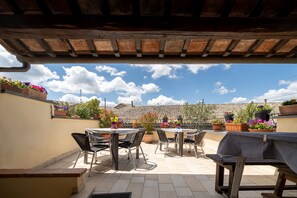 Una veranda esterna con pavimento piastrellato, una panca di legno e un tavolo con sedie. Vasi con piante e fiori adornano le pareti.