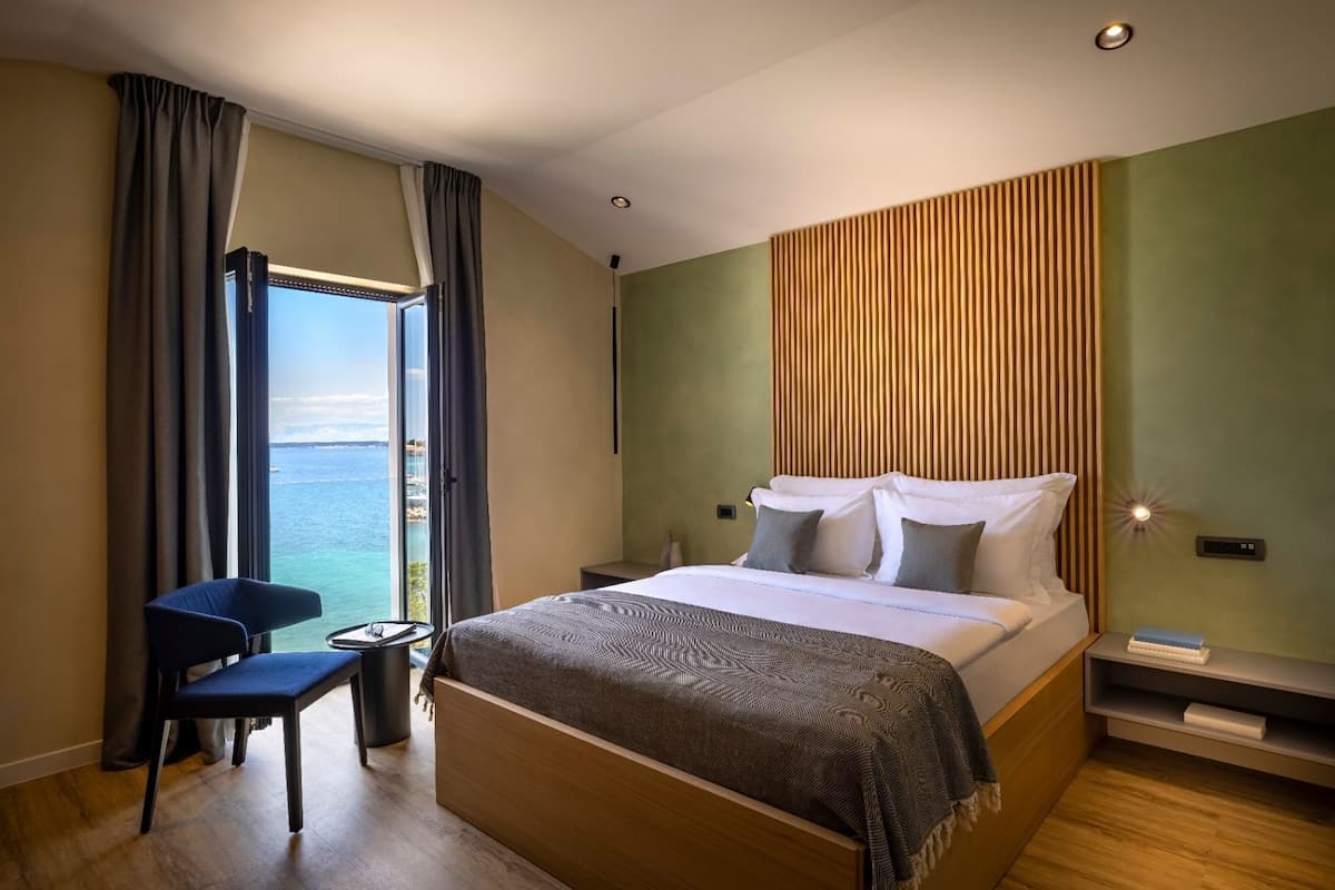 A modern hotel room with a large bed, a chair, a bedside table, and a view of the sea.