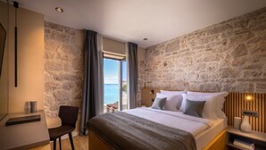 A bedroom with a stone wall, a bed with white linens, a desk with a chair, and a view of the sea. - DUNATOVI DVORI Heritage Hotel (Preko)