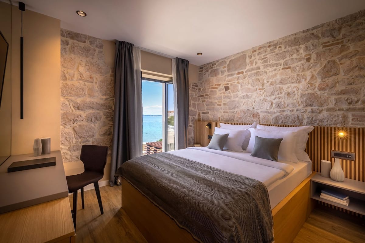 A bedroom with a stone wall, a bed with white bedding, a desk with a chair, and a view of the sea.