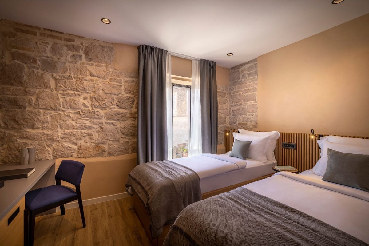 A hotel room with a stone wall, two beds, a desk, and a chair.