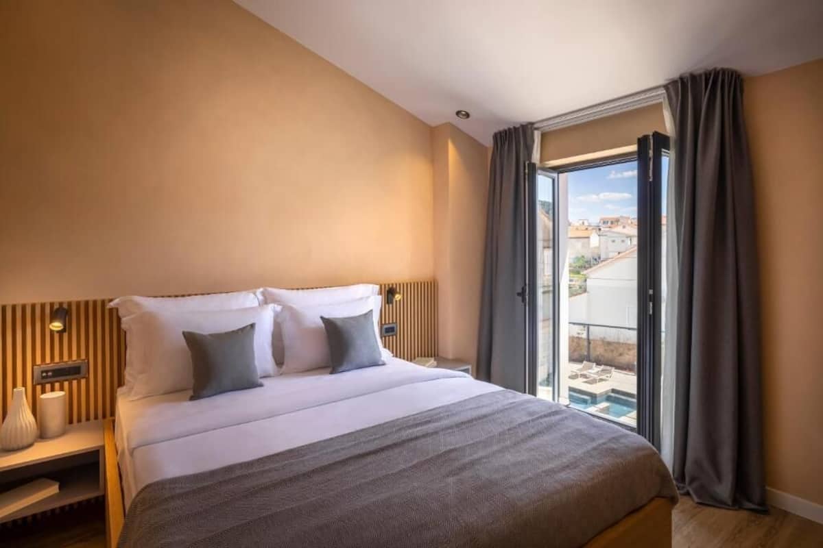 A hotel room with a large bed, a bedside table with a lamp, a window with curtains, and a view of buildings outside.