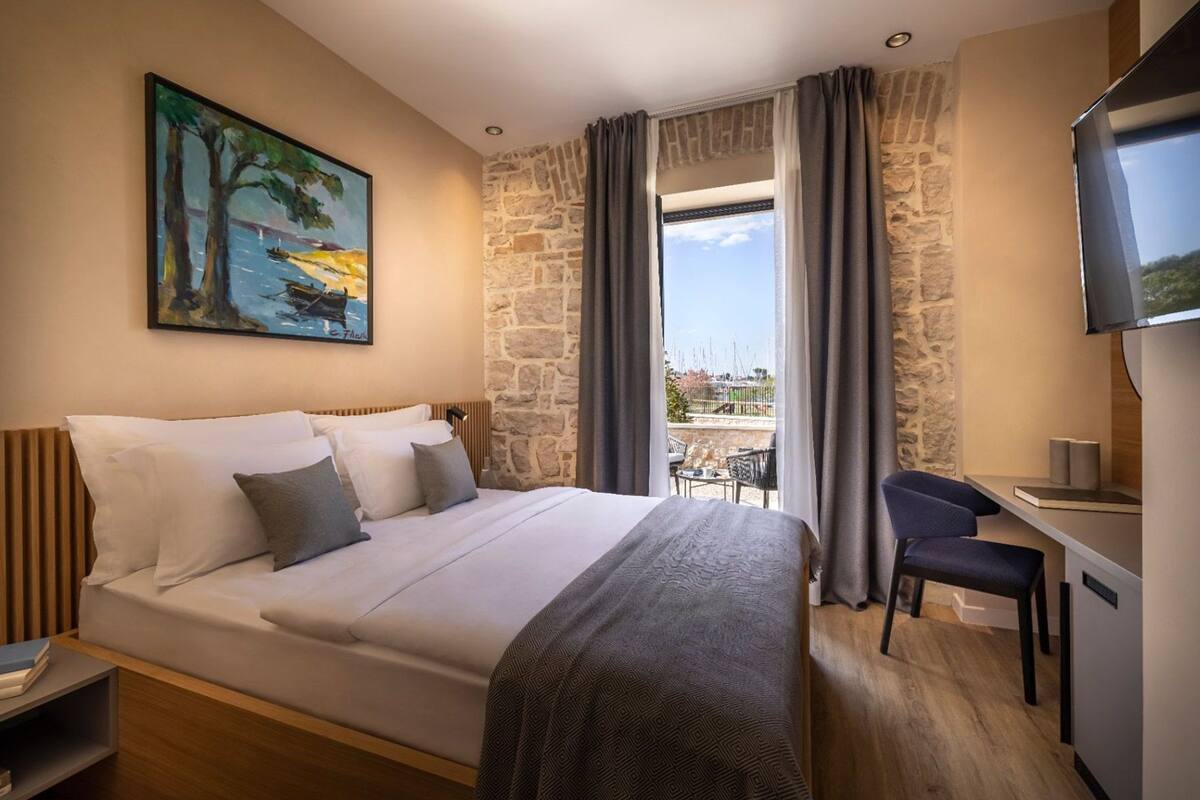 A hotel room with a bed, a desk, a chair, a painting, and a view of the outdoors.
