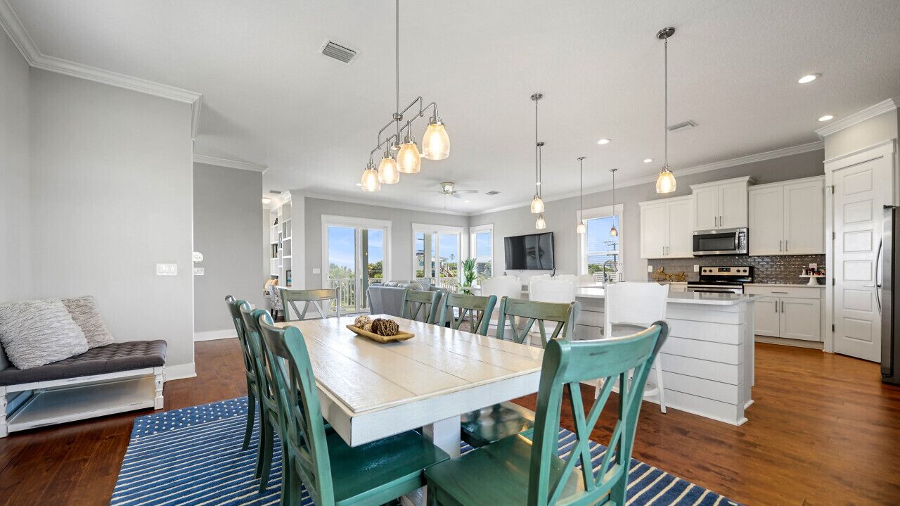 A modern kitchen with white cabinets, a dining area with a wooden table and teal chairs, and a large window providing natural light.