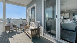 Een overdekt balkon met houten stoelen en een bank, grote schuifdeuren die leiden naar een woonruimte met een bank en een keuken op de achtergrond.